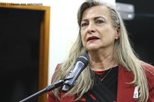 Mulher branca, de cabelos longos e loiros fala em um microfone, ela é Maria Lúcia Fatorelli. Ela está vestida com um paletó vermelho e uma camisa preta.