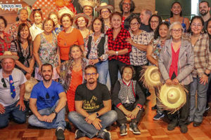 Esta imagem mostra integrantes do Núcleo de Aposentados e Pensionistas do posando para uma foto durante o Arraiá de 18 anos do Núcleo. A maioria está em pé, enquanto algumas estão ajoelhadas ou sentadas na frente. Muitas pessoas estão usando roupas típicas de festa junina, como camisas xadrez, chapéus de palha e acessórios coloridos. O clima é festivo e alegre, com todos sorrindo para a câmera. Ao fundo, há decoração típica de festa junina, como bandeirinhas coloridas e balões.