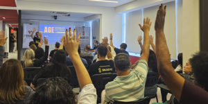 A imagem mostra uma assembleia com servidores do TJMG, muitos deles com a mão levantada, indicando votação. No fundo, um telão exibe o título da AGE (Assembleia Geral Extraordinária) com faixas da FENAJUD e do SINJUS posicionadas ao lado. O texto sobreposto informa que a categoria aprovou uma campanha contra a cultura do atraso do TJMG e elegeu delegados para o Consinjus. O cenário é de engajamento coletivo em uma sala clara e organizada, reforçando a mobilização da categoria por melhorias institucionais.