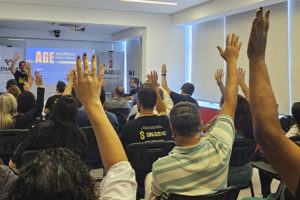 A imagem mostra uma assembleia com servidores do TJMG, muitos deles com a mão levantada, indicando votação. No fundo, um telão exibe o título da AGE (Assembleia Geral Extraordinária) com faixas da FENAJUD e do SINJUS posicionadas ao lado. O texto sobreposto informa que a categoria aprovou uma campanha contra a cultura do atraso do TJMG e elegeu delegados para o Consinjus. O cenário é de engajamento coletivo em uma sala clara e organizada, reforçando a mobilização da categoria por melhorias institucionais.