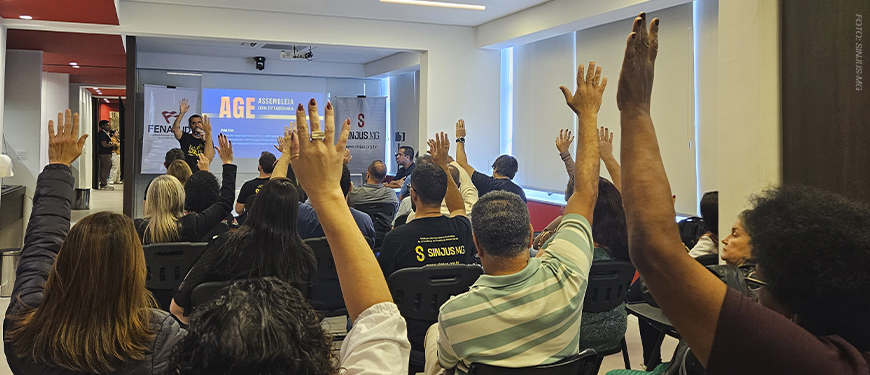 A imagem mostra uma assembleia com servidores do TJMG, muitos deles com a mão levantada, indicando votação. No fundo, um telão exibe o título da AGE (Assembleia Geral Extraordinária) com faixas da FENAJUD e do SINJUS posicionadas ao lado. O texto sobreposto informa que a categoria aprovou uma campanha contra a cultura do atraso do TJMG e elegeu delegados para o Consinjus. O cenário é de engajamento coletivo em uma sala clara e organizada, reforçando a mobilização da categoria por melhorias institucionais.
