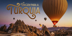 Imagem Acessível: Card de divulgação de viagem para a Turquia, com fundo azul e paisagem da Capadócia ao pôr do sol, marcada por formações rochosas e vários balões coloridos no céu. Em destaque, o texto diz: “Viagem para a Turquia” e “Viva momentos inesquecíveis entre o Oriente e o Ocidente”. A viagem será de 18 a 28 de outubro de 2026, com roteiro por Istambul, Capadócia, Pamukkale, Izmir e Bursa. A peça informa que as vagas são limitadas e que as inscrições devem ser feitas pelo formulário.