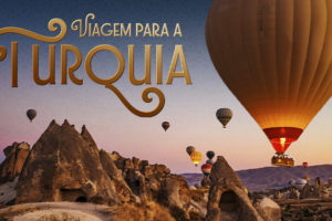 Imagem Acessível: Card de divulgação de viagem para a Turquia, com fundo azul e paisagem da Capadócia ao pôr do sol, marcada por formações rochosas e vários balões coloridos no céu. Em destaque, o texto diz: “Viagem para a Turquia” e “Viva momentos inesquecíveis entre o Oriente e o Ocidente”. A viagem será de 18 a 28 de outubro de 2026, com roteiro por Istambul, Capadócia, Pamukkale, Izmir e Bursa. A peça informa que as vagas são limitadas e que as inscrições devem ser feitas pelo formulário.