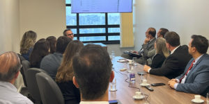 Imagem Acessível: A imagem mostra uma sala de reuniões com várias pessoas sentadas ao redor de uma mesa, observando uma tela que exibe dados técnicos. O texto sobreposto diz: “Sindicatos discutem necessidade de rever critérios do cálculo da força de trabalho”.