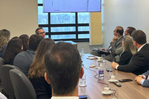 Imagem Acessível: A imagem mostra uma sala de reuniões com várias pessoas sentadas ao redor de uma mesa, observando uma tela que exibe dados técnicos. O texto sobreposto diz: “Sindicatos discutem necessidade de rever critérios do cálculo da força de trabalho”.
