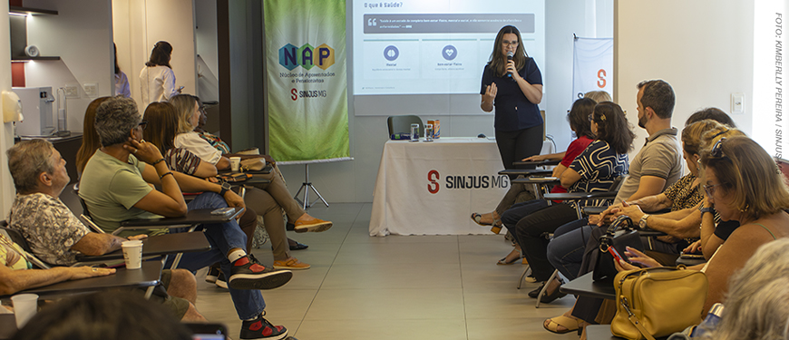 Imagem acessível: A imagem mostra aposentados e pensionistas reunidos em uma palestra promovida pelo NAP do SINJUS-MG, enquanto uma fisioterapeuta conduz a apresentação. O encontro teve como tema “Viver bem é uma questão de equilíbrio” e abordou a importância do equilíbrio físico, emocional e social para o bem-estar e a autonomia na terceira idade.