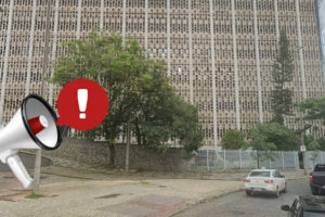 Foto diurna da fachada lateral do edifício situado à Praça Milton Campos, feita de baixo para cima. Sobre ela, é aplicada a figura de um megafone branco, vermelho e preto, do qual sai um balão vermelho com uma exclamação em branco, indicando atenção.