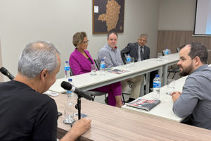 eunião em uma sala de conferência, com seis pessoas sentadas em torno de mesas em formato de U. Dois homens à esquerda falam ao microfone, enquanto os demais participantes — entre eles uma mulher de blazer roxo e quatro homens — ouvem atentamente. Há garrafas de água e revistas sobre a mesa. Ao fundo, um telão exibe a identidade visual do projeto 'Se Liga, Minas'.