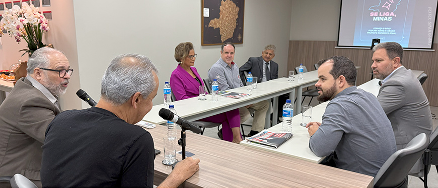 eunião em uma sala de conferência, com seis pessoas sentadas em torno de mesas em formato de U. Dois homens à esquerda falam ao microfone, enquanto os demais participantes — entre eles uma mulher de blazer roxo e quatro homens — ouvem atentamente. Há garrafas de água e revistas sobre a mesa. Ao fundo, um telão exibe a identidade visual do projeto 'Se Liga, Minas'.