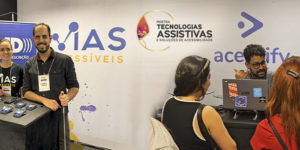 Montagem com duas fotos do estande da Vias Acessíveis na Mostra de Tecnologias Assistivas e Soluções de Acessibilidade. Na parte superior, uma mulher e um homem posam sorrindo atrás de uma bancada com materiais e equipamentos de acessibilidade, diante do painel da empresa. Na parte inferior, um expositor conversa com duas mulheres em frente a um notebook, enquanto apresenta soluções e recursos disponíveis no estande. Sobre as imagens, aparece a frase: “Inclusão efetiva de pessoas com deficiência exige acessibilidade desde a concepção dos eventos”. A Mostra reuniu empresas, universidades e empreendedores para apresentar tecnologias e soluções voltadas à acessibilidade e à inclusão.