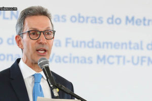 Governador de Minas Gerais, Romeu Zema (homem de pele clara, cabelos grisalhos curtos e usando óculos preto e camisa cinza) em pé, durante pronunciamento em um púlpito e segurando uma folha com o seu texto de discurso.