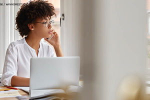 Imagem Acessível: Mulher negra está em uma chamada de voz, enquanto trabalha diante de um notebook em seu home office. Ela está em um ambiente claro e amplo, olhando para a janela de onde se avistam outros prédios. Conteúdo textual: ATÉ 31/1 - Últimos dias para gestores responderem pesquisa sobre teletrabalho.