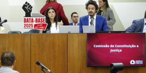 A imagem mostra uma sessão da Comissão de Constituição e Justiça (CCJ) da Assembleia Legislativa de Minas Gerais, com o parlamentar Doorgal Andrada na presidência da Comissão, falando ao microfone. Há ainda um selo com o punho erguido e a frase “Data-Base 2025 – Eu não abro mão!”, reforçando o tom de pressão e mobilização da categoria pela aprovação do projeto.