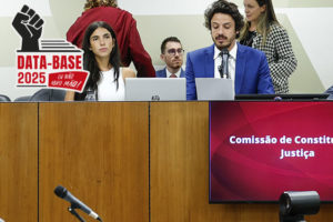 A imagem mostra uma sessão da Comissão de Constituição e Justiça (CCJ) da Assembleia Legislativa de Minas Gerais, com o parlamentar Doorgal Andrada na presidência da Comissão, falando ao microfone. Há ainda um selo com o punho erguido e a frase “Data-Base 2025 – Eu não abro mão!”, reforçando o tom de pressão e mobilização da categoria pela aprovação do projeto.