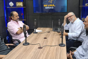 Num cenário em tons de azul há uma mesa com microfones onde estão 4 pessoas: o anfitrião do podcast Se liga Minas, Orion Teixeira; o ex-prefeito de Belo Horizonte, Alexandre Kalil; e os dirigentes do SINJUS-MG, Felipe Rodrigues (diretor de Assuntos Jurídicos) e Alex Aguiar (diretor de Finanças).