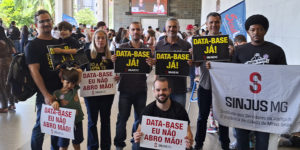A imagem mostra um grupo de servidoras e servidores e dirigentes do SINJUS-MG reunidos na ALMG, segurando cartazes com frases como “DATA-BASE JÁ!” e “DATA-BASE EU NÃO ABRO MÃO!”, além de uma bandeira do SINJUS MG – Sindicato dos Servidores da Justiça de 2ª Instância do Estado de Minas Gerais. No topo, aparece a versal “RECOMPOSIÇÃO” e, em destaque, o texto: “Com forte mobilização sindical, Data-Base 2025 é aprovada em definitivo na ALMG”.