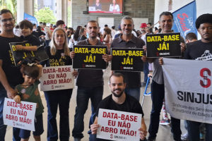 A imagem mostra um grupo de servidoras e servidores e dirigentes do SINJUS-MG reunidos na ALMG, segurando cartazes com frases como “DATA-BASE JÁ!” e “DATA-BASE EU NÃO ABRO MÃO!”, além de uma bandeira do SINJUS MG – Sindicato dos Servidores da Justiça de 2ª Instância do Estado de Minas Gerais. No topo, aparece a versal “RECOMPOSIÇÃO” e, em destaque, o texto: “Com forte mobilização sindical, Data-Base 2025 é aprovada em definitivo na ALMG”.