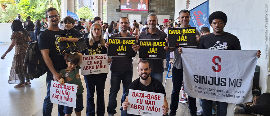 A imagem mostra um grupo de servidoras e servidores e dirigentes do SINJUS-MG reunidos na ALMG, segurando cartazes com frases como “DATA-BASE JÁ!” e “DATA-BASE EU NÃO ABRO MÃO!”, além de uma bandeira do SINJUS MG – Sindicato dos Servidores da Justiça de 2ª Instância do Estado de Minas Gerais. No topo, aparece a versal “RECOMPOSIÇÃO” e, em destaque, o texto: “Com forte mobilização sindical, Data-Base 2025 é aprovada em definitivo na ALMG”.