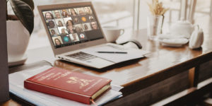 : A imagem de fundo mostra um notebook sobre uma mesa, exibindo uma reunião virtual com várias mulheres na tela, representando o encontro on-line. Ao lado, há uma xícara, bloco de anotações e caneta, compondo ambiente de estudo e reflexão. Em primeiro plano, aparece o livro sobre a mesa “Mulheres que correm com os lobos”, de Clarissa Pinkola Estés, reforçando o tema do debate. No topo, aparece escrito a versal “CLUBE DE LEITURA” e, em destaque logo abaixo, o texto: “NM se reúne para debater a reconexão com a essência feminina”.