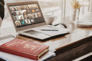 : A imagem de fundo mostra um notebook sobre uma mesa, exibindo uma reunião virtual com várias mulheres na tela, representando o encontro on-line. Ao lado, há uma xícara, bloco de anotações e caneta, compondo ambiente de estudo e reflexão. Em primeiro plano, aparece o livro sobre a mesa “Mulheres que correm com os lobos”, de Clarissa Pinkola Estés, reforçando o tema do debate. No topo, aparece escrito a versal “CLUBE DE LEITURA” e, em destaque logo abaixo, o texto: “NM se reúne para debater a reconexão com a essência feminina”.