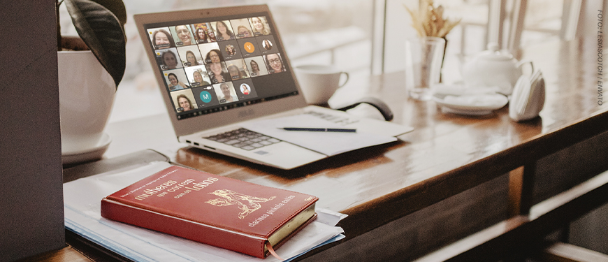 : A imagem de fundo mostra um notebook sobre uma mesa, exibindo uma reunião virtual com várias mulheres na tela, representando o encontro on-line. Ao lado, há uma xícara, bloco de anotações e caneta, compondo ambiente de estudo e reflexão. Em primeiro plano, aparece o livro sobre a mesa “Mulheres que correm com os lobos”, de Clarissa Pinkola Estés, reforçando o tema do debate. No topo, aparece escrito a versal “CLUBE DE LEITURA” e, em destaque logo abaixo, o texto: “NM se reúne para debater a reconexão com a essência feminina”.
