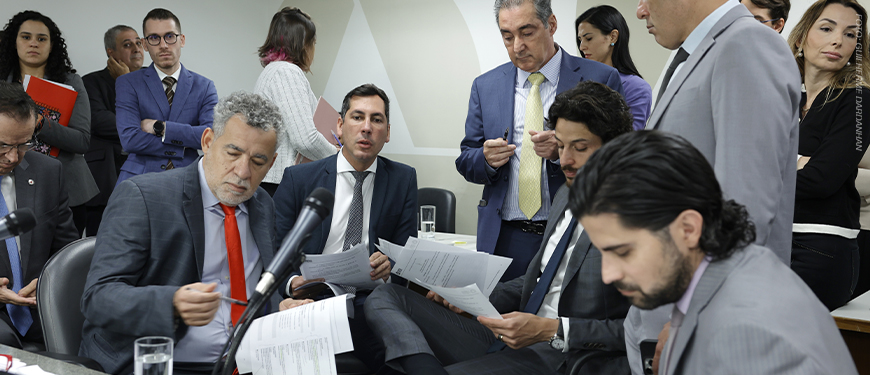 A imagem mostra um grupo de parlamentares reunidos em uma sala da ALMG, discutindo e analisando documentos. O ambiente é formal, com todos trajando ternos. O texto sobre a imagem informa que o Propag começou a tramitar na ALMG e que o SINJUS está intensificando articulações políticas em várias esferas, reforçando sua atuação na defesa dos direitos das servidoras e servidores mineiros