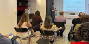 Registro de uma reunião realizada no Espaço Lea Leda do SINJUS-MG. Na imagem, o coordenador-geral Felipe Rodrigues fala ao microfone diante de uma tela que exibe a minuta de regulamentação da jornada, enquanto servidoras e servidores acompanham a apresentação.