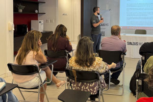 Registro de uma reunião realizada no Espaço Lea Leda do SINJUS-MG. Na imagem, o coordenador-geral Felipe Rodrigues fala ao microfone diante de uma tela que exibe a minuta de regulamentação da jornada, enquanto servidoras e servidores acompanham a apresentação.