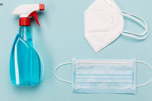 Imagem acessível: Sobre uma superfície plana azul e limpa estão um spray de álcool em gel, uma máscara cirúrgica de tripla camada e uma máscara KN95. Conteúdo textual: COVID-1+9 - Após aumento de casos, TJ reforça medidas de prevenção