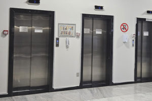 A imagem mostra o corredor interno do Anexo 1 com três elevadores lado a lado que apresentam problemas. Acima de cada elevador há um painel digital indicando o andar. Entre os elevadores, há placas informativas e um telefone de emergência. À direita, vê-se um dispensador de álcool em gel e uma placa proibindo o uso de celular.
