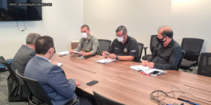 Em uma sala de reuniões estão assentados em uma grande mesa de madeira 5 homens de pele clara utilizando máscaras e vestindo roupa social.