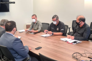 Em uma sala de reuniões estão assentados em uma grande mesa de madeira 5 homens de pele clara utilizando máscaras e vestindo roupa social.