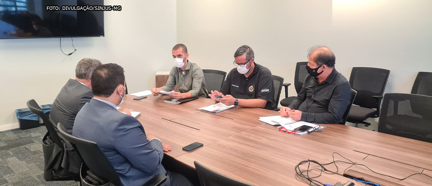 Em uma sala de reuniões estão assentados em uma grande mesa de madeira 5 homens de pele clara utilizando máscaras e vestindo roupa social.