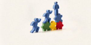 Imagem Acessível: Imagem com fundo neutro em tom claro. Na parte inferior da imagem, há uma ilustração com três figuras humanas estilizadas na cor azul, representadas de forma simples e sem detalhes faciais. Elas estão posicionadas sobre peças de quebra-cabeça coloridas (verde, amarelo, vermelho e azul), formando uma escada. Uma das figuras está no nível mais alto, em posição ereta; outra está subindo, com uma perna apoiada em uma peça mais elevada; e a terceira está em posição inicial, sugerindo movimento de ascensão. A composição transmite ideia de progresso, crescimento profissional e avanço na carreira. Conteúdo textual: CARREIRA - TJMG divulga lista definitiva de inscritos e de inscrições não conhecidas da PV 2024