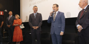 Imagem Acessível: Registro de uma sessão de cinema. Em frente à tela de projeção, estão quatro pessoas. À direita está o presidente do TJMG, Luiz Carlos Corrêa Junior, um homem de meia-idade, com pele clara, usando terno azul e gravata listrada, segura um microfone e fala ao público, com expressão segura e comunicativa. Ao centro, está Alex Aguiar, diretor de Finanças do SINUJUS-MG e coordenador do Núcleo da Pessoa com Deficiência do Sindicato. Ele também aparenta ter meia-idade, tem pele morena e cabelos curtos grisalhos, veste terno escuro e observa o orador com um leve sorriso. À esquerda, está a desembargadora Ana Conceição Barbuda Ferreira do Tribunal da Justiça da Bahia, uma mulher de pele clara, com cabelos curtos e usa óculos. Ela usa um vestido laranja e segura uma bolsa clara, sorrindo. Ao lado dela, está Wagner Ferreira, vereador de BH e diretor de Formação Política e Sindical do SINJUS, ele é um homem negro, tem os cabelos grisalhos e veste um terno escuro. Ele também demonstra atenção à fala do presidente do TJMG. Na parte inferior da imagem, vê-se o público sentado em poltronas vermelhas, assistindo à apresentação. Na tela ao fundo, aparece um texto projetado, parcialmente legível: “Aguarde, a sessão já vai começar”, junto à palavra “EXISTO”. Conteúdo textual: EXISTO - SINJUS lança documentário sobre luta das pessoas com deficiência.