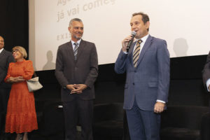 Imagem Acessível: Registro de uma sessão de cinema. Em frente à tela de projeção, estão quatro pessoas. À direita está o presidente do TJMG, Luiz Carlos Corrêa Junior, um homem de meia-idade, com pele clara, usando terno azul e gravata listrada, segura um microfone e fala ao público, com expressão segura e comunicativa. Ao centro, está Alex Aguiar, diretor de Finanças do SINUJUS-MG e coordenador do Núcleo da Pessoa com Deficiência do Sindicato. Ele também aparenta ter meia-idade, tem pele morena e cabelos curtos grisalhos, veste terno escuro e observa o orador com um leve sorriso. À esquerda, está a desembargadora Ana Conceição Barbuda Ferreira do Tribunal da Justiça da Bahia, uma mulher de pele clara, com cabelos curtos e usa óculos. Ela usa um vestido laranja e segura uma bolsa clara, sorrindo. Ao lado dela, está Wagner Ferreira, vereador de BH e diretor de Formação Política e Sindical do SINJUS, ele é um homem negro, tem os cabelos grisalhos e veste um terno escuro. Ele também demonstra atenção à fala do presidente do TJMG. Na parte inferior da imagem, vê-se o público sentado em poltronas vermelhas, assistindo à apresentação. Na tela ao fundo, aparece um texto projetado, parcialmente legível: “Aguarde, a sessão já vai começar”, junto à palavra “EXISTO”. Conteúdo textual: EXISTO - SINJUS lança documentário sobre luta das pessoas com deficiência.