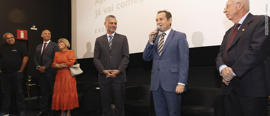 Imagem Acessível: Registro de uma sessão de cinema. Em frente à tela de projeção, estão quatro pessoas. À direita está o presidente do TJMG, Luiz Carlos Corrêa Junior, um homem de meia-idade, com pele clara, usando terno azul e gravata listrada, segura um microfone e fala ao público, com expressão segura e comunicativa. Ao centro, está Alex Aguiar, diretor de Finanças do SINUJUS-MG e coordenador do Núcleo da Pessoa com Deficiência do Sindicato. Ele também aparenta ter meia-idade, tem pele morena e cabelos curtos grisalhos, veste terno escuro e observa o orador com um leve sorriso. À esquerda, está a desembargadora Ana Conceição Barbuda Ferreira do Tribunal da Justiça da Bahia, uma mulher de pele clara, com cabelos curtos e usa óculos. Ela usa um vestido laranja e segura uma bolsa clara, sorrindo. Ao lado dela, está Wagner Ferreira, vereador de BH e diretor de Formação Política e Sindical do SINJUS, ele é um homem negro, tem os cabelos grisalhos e veste um terno escuro. Ele também demonstra atenção à fala do presidente do TJMG. Na parte inferior da imagem, vê-se o público sentado em poltronas vermelhas, assistindo à apresentação. Na tela ao fundo, aparece um texto projetado, parcialmente legível: “Aguarde, a sessão já vai começar”, junto à palavra “EXISTO”. Conteúdo textual: EXISTO - SINJUS lança documentário sobre luta das pessoas com deficiência.