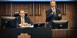 À esquerda, está outro homem, o presidente do TJMG, desembargador Luiz Carlos Corrêa Junior, ele também aparenta meia-idade, tem pele clara e cabelos escuros, está sentado atrás de uma bancada elevada. Ele também veste uma toga sobre um terno escuro, camisa clara e gravata, e observa atentamente o orador, com expressão séria. À frente da bancada, há um brasão em destaque. Sobre a mesa, estão equipamentos como monitor, microfone e documentos. O fundo é composto por painéis de madeira com acabamento vertical, conferindo um aspecto solene e formal ao espaço.