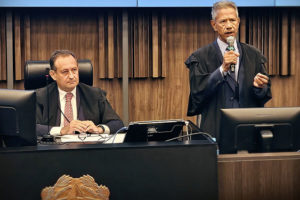 À esquerda, está outro homem, o presidente do TJMG, desembargador Luiz Carlos Corrêa Junior, ele também aparenta meia-idade, tem pele clara e cabelos escuros, está sentado atrás de uma bancada elevada. Ele também veste uma toga sobre um terno escuro, camisa clara e gravata, e observa atentamente o orador, com expressão séria. À frente da bancada, há um brasão em destaque. Sobre a mesa, estão equipamentos como monitor, microfone e documentos. O fundo é composto por painéis de madeira com acabamento vertical, conferindo um aspecto solene e formal ao espaço.