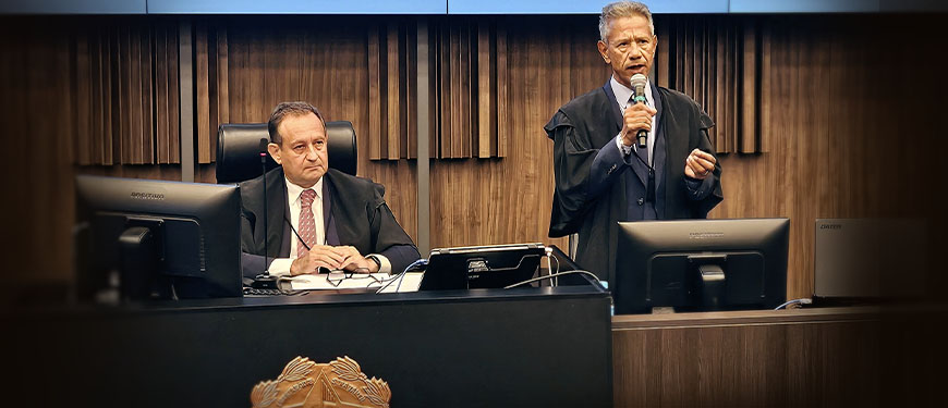 À esquerda, está outro homem, o presidente do TJMG, desembargador Luiz Carlos Corrêa Junior, ele também aparenta meia-idade, tem pele clara e cabelos escuros, está sentado atrás de uma bancada elevada. Ele também veste uma toga sobre um terno escuro, camisa clara e gravata, e observa atentamente o orador, com expressão séria. À frente da bancada, há um brasão em destaque. Sobre a mesa, estão equipamentos como monitor, microfone e documentos. O fundo é composto por painéis de madeira com acabamento vertical, conferindo um aspecto solene e formal ao espaço.