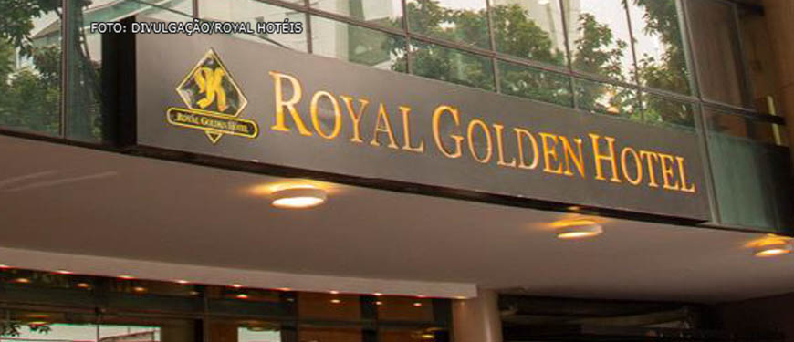 Fachada de edifício com vidros espelhados em tom dourado. Há uma placa com fundo preto, onde sê lê "Royal Golden Hotel" em amarelo.