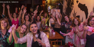 Foto noturna em um ambiente aconchegante com servidoras que participam do Núcleo da Mulher, as diretoras Adriana Gonçalves Mota Teodoro e Janaína Torres Barbosa e a comediante Paloma Santos o semblante é de alegria e festa entre elas.