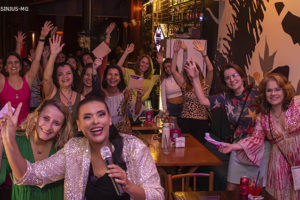 Foto noturna em um ambiente aconchegante com servidoras que participam do Núcleo da Mulher, as diretoras Adriana Gonçalves Mota Teodoro e Janaína Torres Barbosa e a comediante Paloma Santos o semblante é de alegria e festa entre elas.
