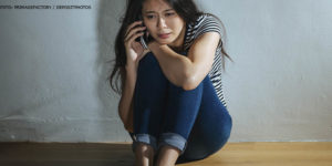 Imagem Acessível: Mulher sentada no chão com postura e expressão facial de medo, ela está falando ao telefone.