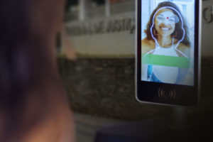 Imagem Acessível: A imagem mostra um terminal de reconhecimento facial em primeiro plano, com a fachada do Tribunal de Justiça de Minas Gerais ao fundo. Conteúdo textual: Faça seu cadastro - TJMG implanta reconhecimento facial para acesso ao Edifício-Sede