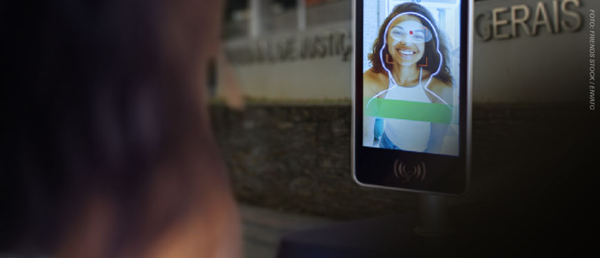 Imagem Acessível: A imagem mostra um terminal de reconhecimento facial em primeiro plano, com a fachada do Tribunal de Justiça de Minas Gerais ao fundo. Conteúdo textual: Faça seu cadastro - TJMG implanta reconhecimento facial para acesso ao Edifício-Sede