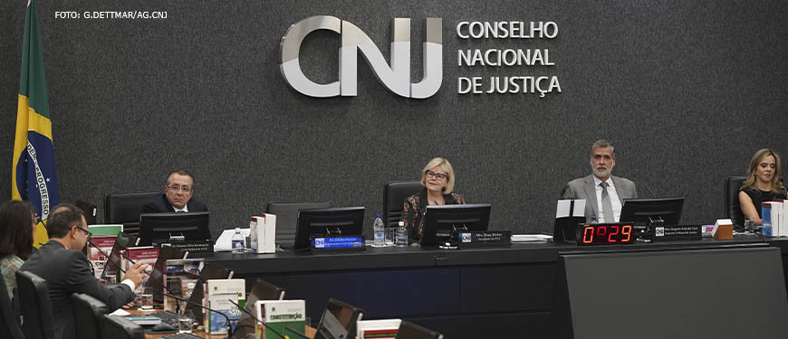 Mesa diretiva do CNJ (Conselho Nacional de Justiça), ao centro está a presidente do CNJ, Ministra Rosa Weber, ela é uma mulher de pele branca, tem cabelos loiros, lisos e curtos; ela veste um traje social escuro estampado e usa óculos de grau.