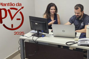 A imagem mostra uma mulher de cabelos castanhos compridos e um homem branco, de barba e cabelos castanhos, vestindo camisa cinza do SINJUS, sentados lado a lado em uma mesa de atendimento, analisando informações em um notebook. O homem que faz o atendimento é o coordenador-geral do Sindicato, Felipe Rodrigues. Sobre a fotografia, aparece o símbolo vermelho do Plantão da PV 2025, com uma seta ascendente integrada a um relógio.