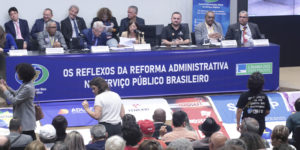 A imagem mostra a audiência pública realizada na Câmara dos Deputados, em Brasília. Na mesa principal, representantes de entidades sindicais e parlamentares discutem. Sobre a imagem, o texto diz: “Fenajud reforça resistência nacional à Reforma Administrativa na Câmara dos Deputados”.