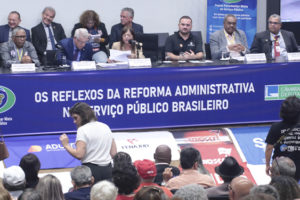 A imagem mostra a audiência pública realizada na Câmara dos Deputados, em Brasília. Na mesa principal, representantes de entidades sindicais e parlamentares discutem. Sobre a imagem, o texto diz: “Fenajud reforça resistência nacional à Reforma Administrativa na Câmara dos Deputados”.